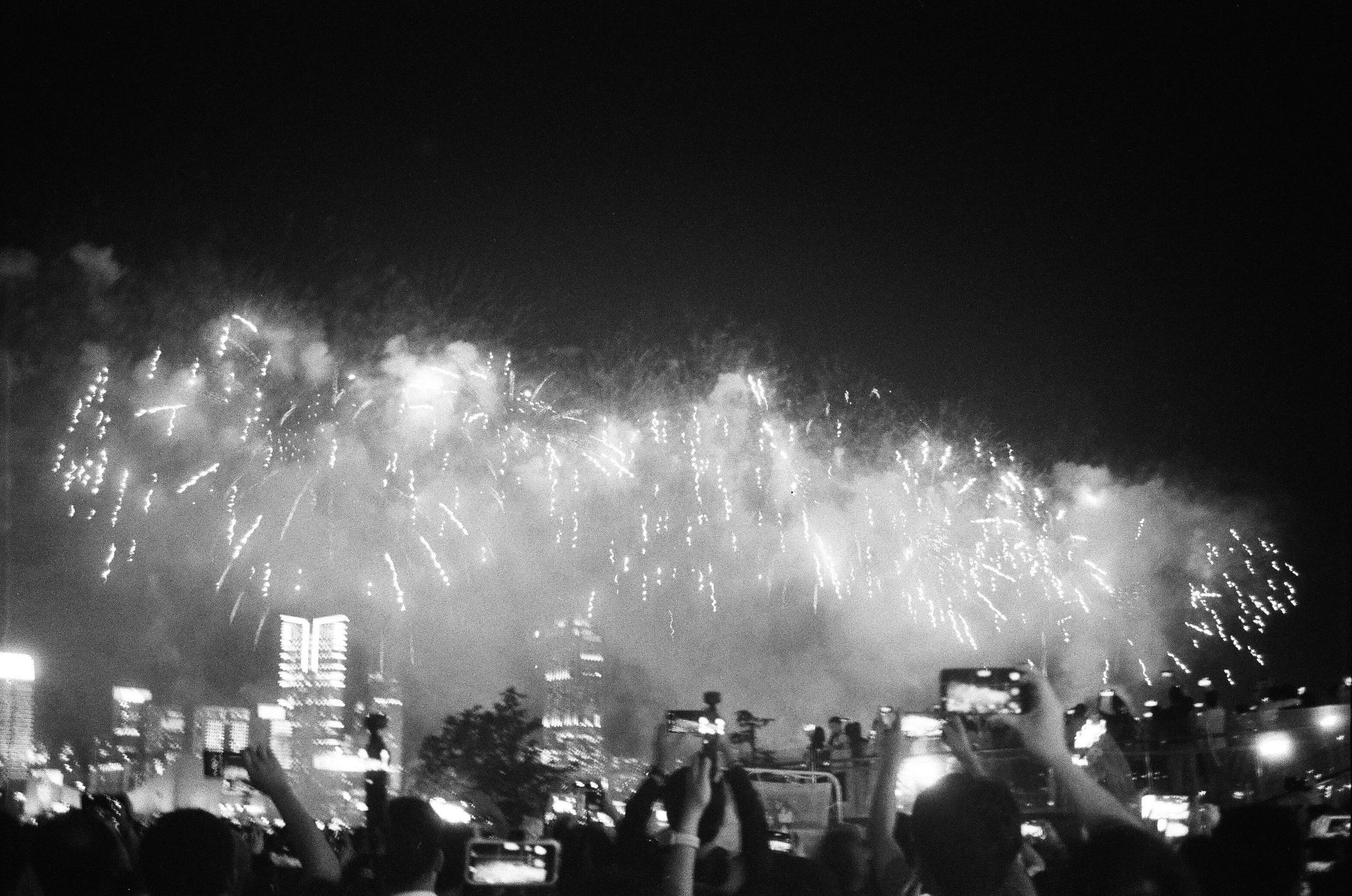 A crowd filming fireworks over a city skyline at night