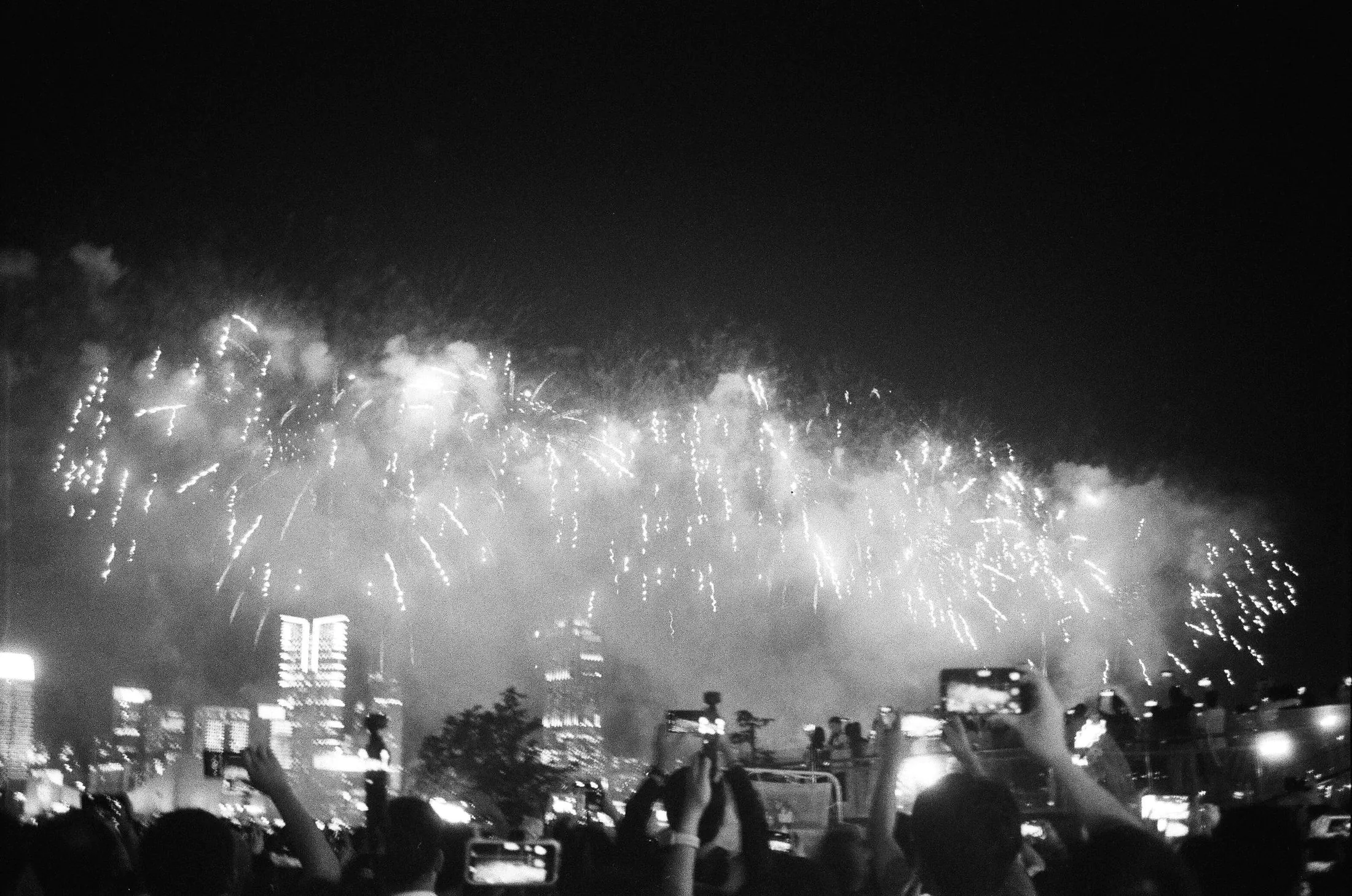 A crowd filming fireworks over a city skyline at night